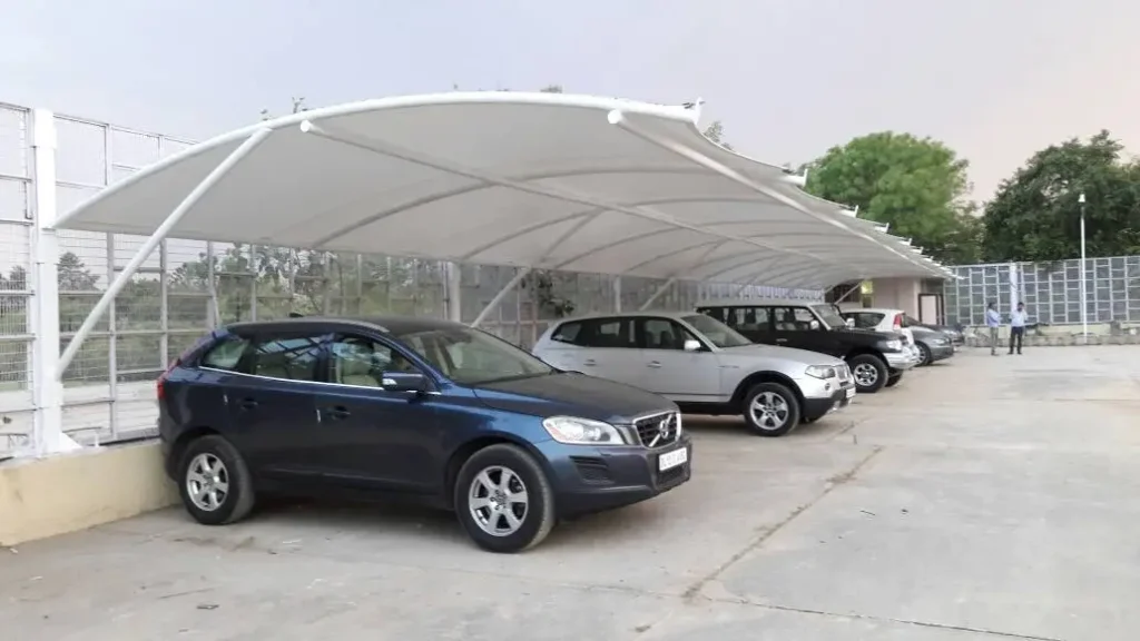 IMG-20191202-WA0004 Row of parked cars under a large white curved shade canopy in a parking lot, with a blue SUV closest to the camera and several other vehicles in the background.