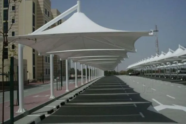 IMG-20190913-WA0009 White tensile shade sails line a pedestrian walkway beside a parking area on a sunny day, creating a long, covered promenade.