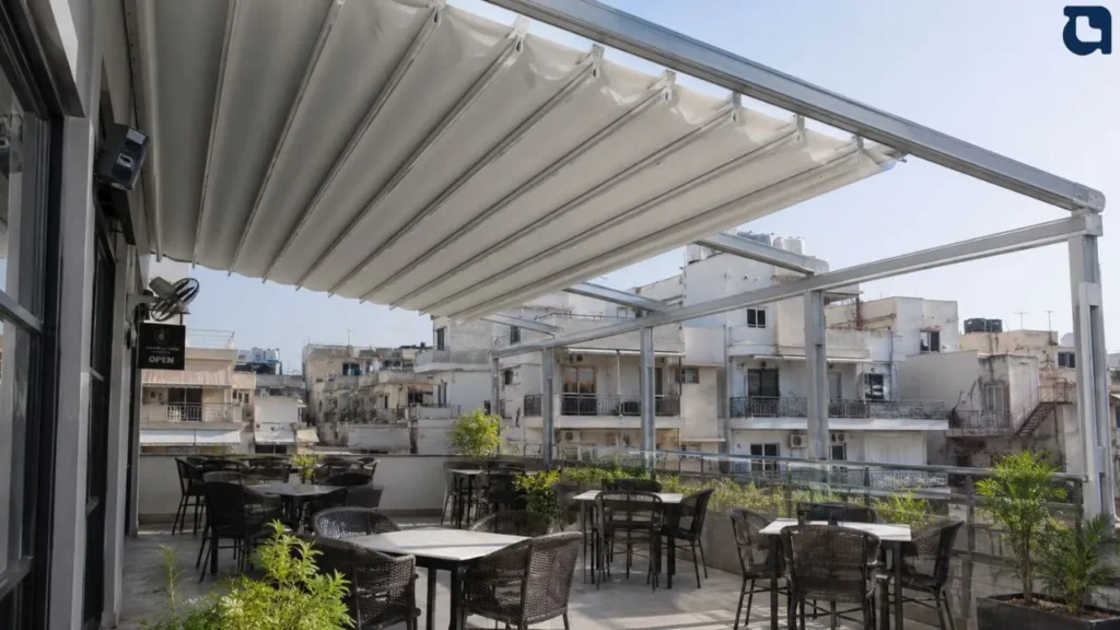 Rooftop café with black wicker chairs and white tables under a white fabric shade, overlooking city buildings.
