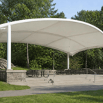 White curved outdoor pavilion with a shade canopy and multiple picnic tables in a park setting.